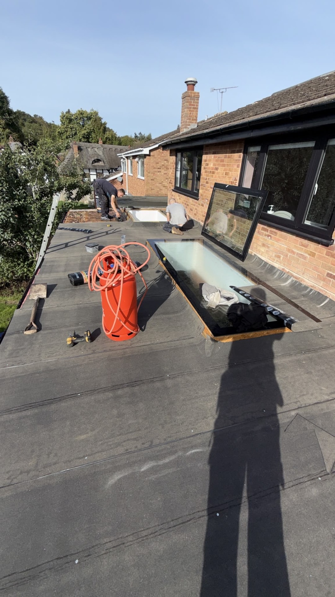 Roofers laying new felt layers across the flat roof, with a gas bottle, tools and an open rooflight on the deck